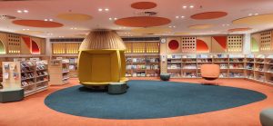 Photograph taken inside the Parramatta Library. Along the back wall are several wooden bookshelves. In the centre, a wooden hive-shaped seat sits on a forest green rug and orange carpet.