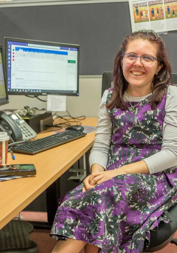 Nikki Andersen is sitting at a desk smiling, wearing a purple floral dress on Wear It Purple Day.
